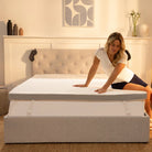 [all] Woman sitting on a Hybrid Mattress Topper in a cozy bedroom setting with neutral decor.