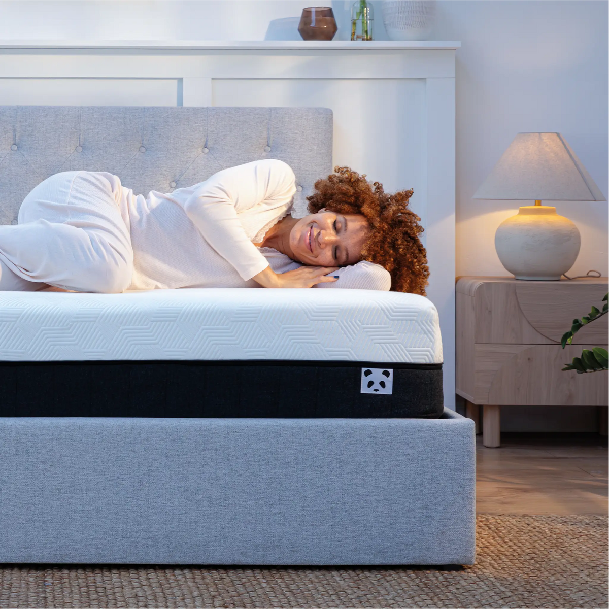 [HybridMattressPro]  A woman in white pajamas lies on her side on a mattress atop a gray upholstered bed frame in a softly lit bedroom. The mattress has a white top layer with a subtle geometric pattern and a black center band, with a small Panda Mattress logo patch visible on the side.